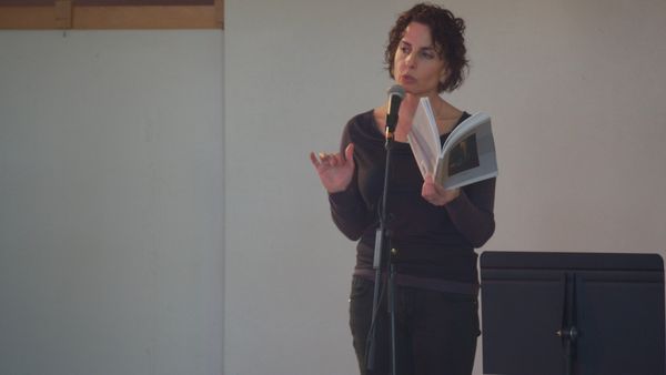 New Mexico Poet Laureate Lauren Camp stands at a microphone, reading from an open book of poetry.